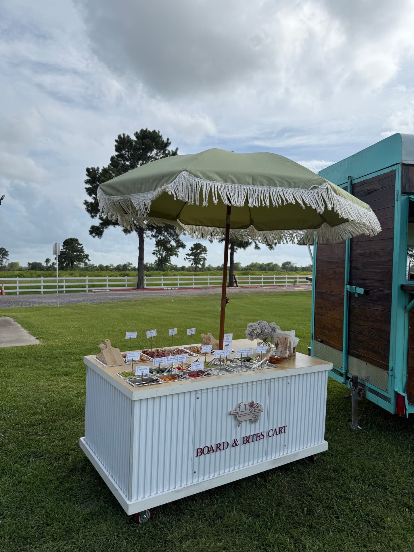 The Board & Bites Cart outdoors on a lawn under its green fringed umbrella, stocked with grazing trays