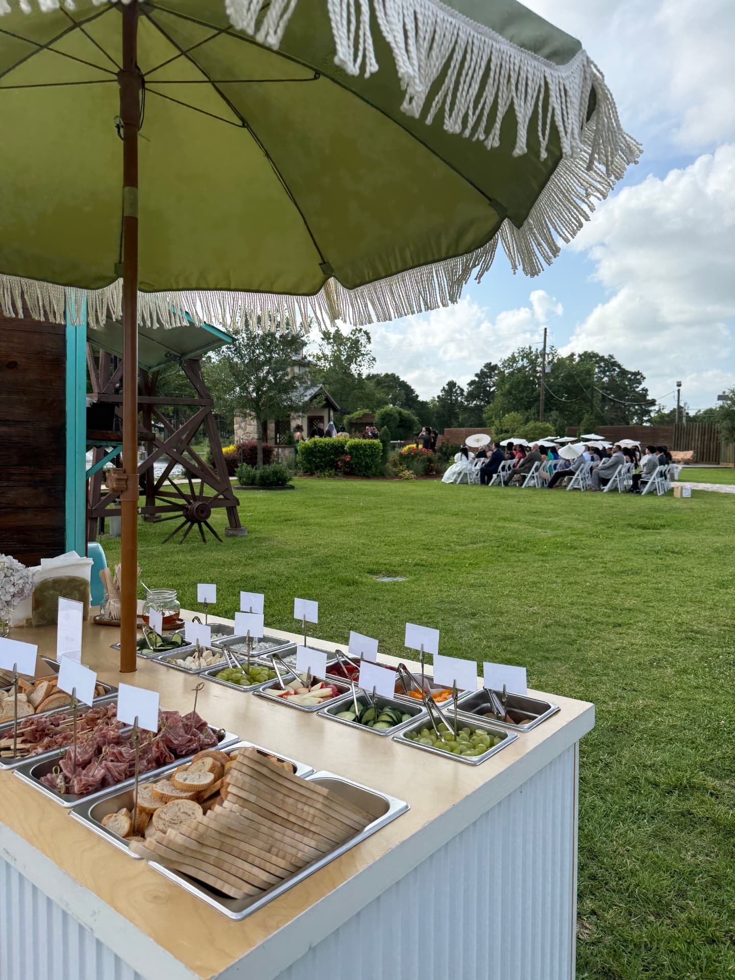 The grazing cart under a fringed umbrella beside an outdoor wedding ceremony